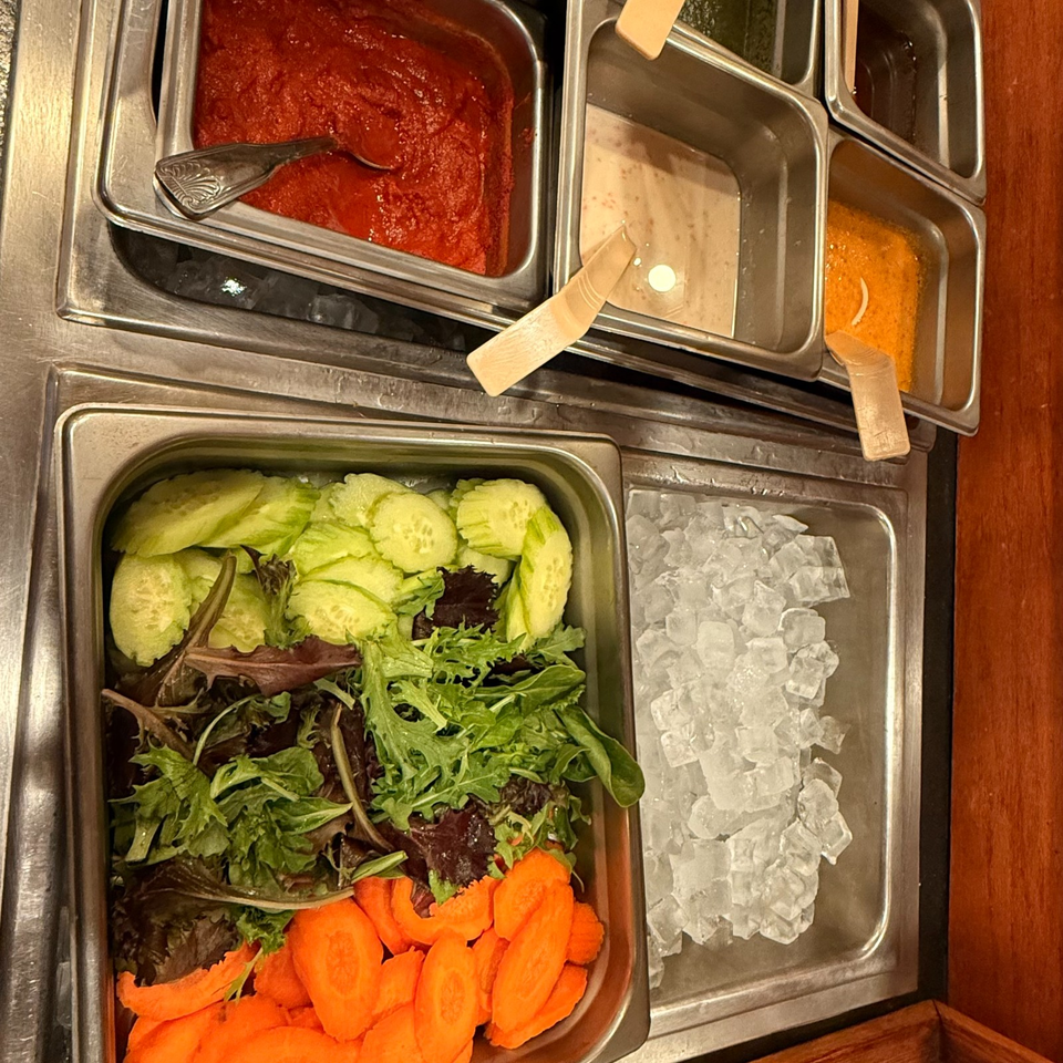 Buffet station with salad greens, sliced cucumbers and carrots on ice, alongside pans of red, creamy, and orange sauces.
