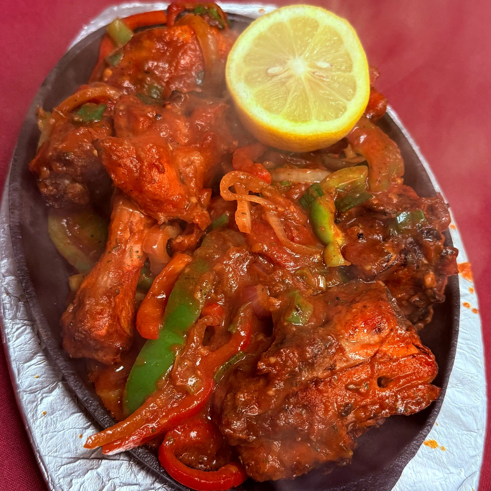 Sizzling platter of saucy grilled chicken with peppers, onions, herbs, and a lemon half.