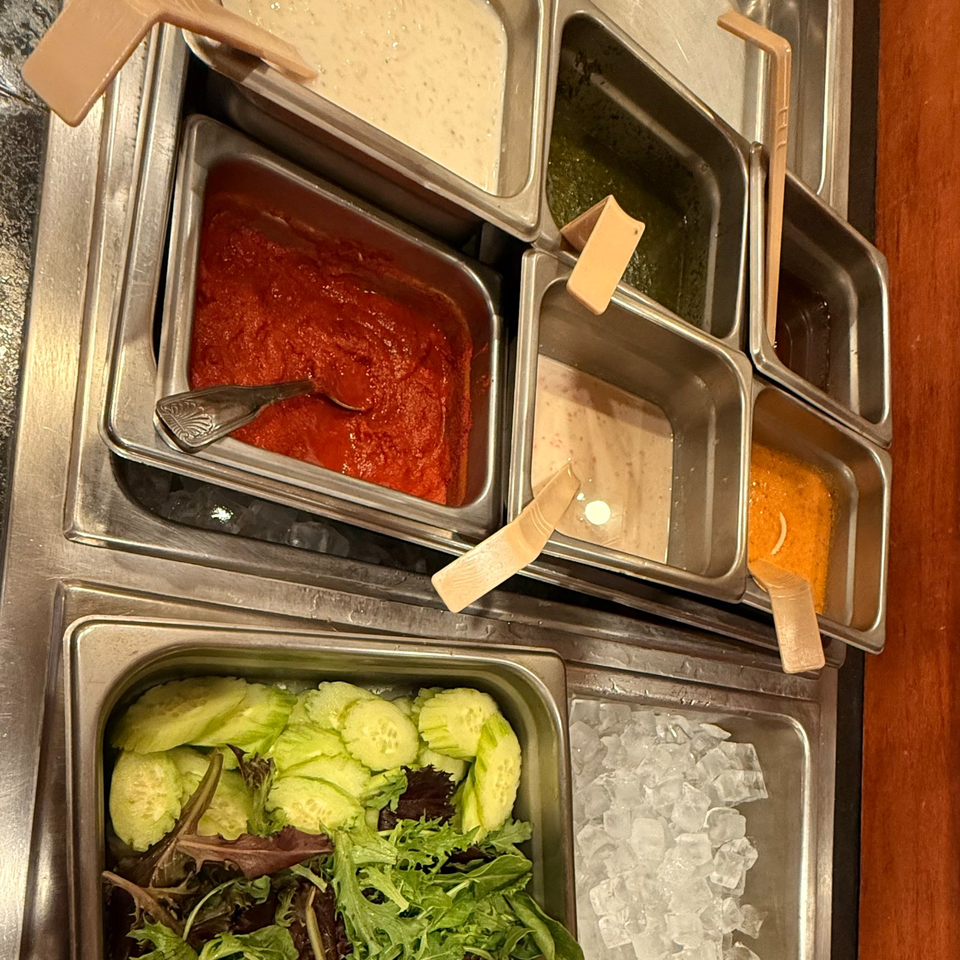Buffet station with trays of colorful sauces, sliced cucumbers, mixed greens, and crushed ice.