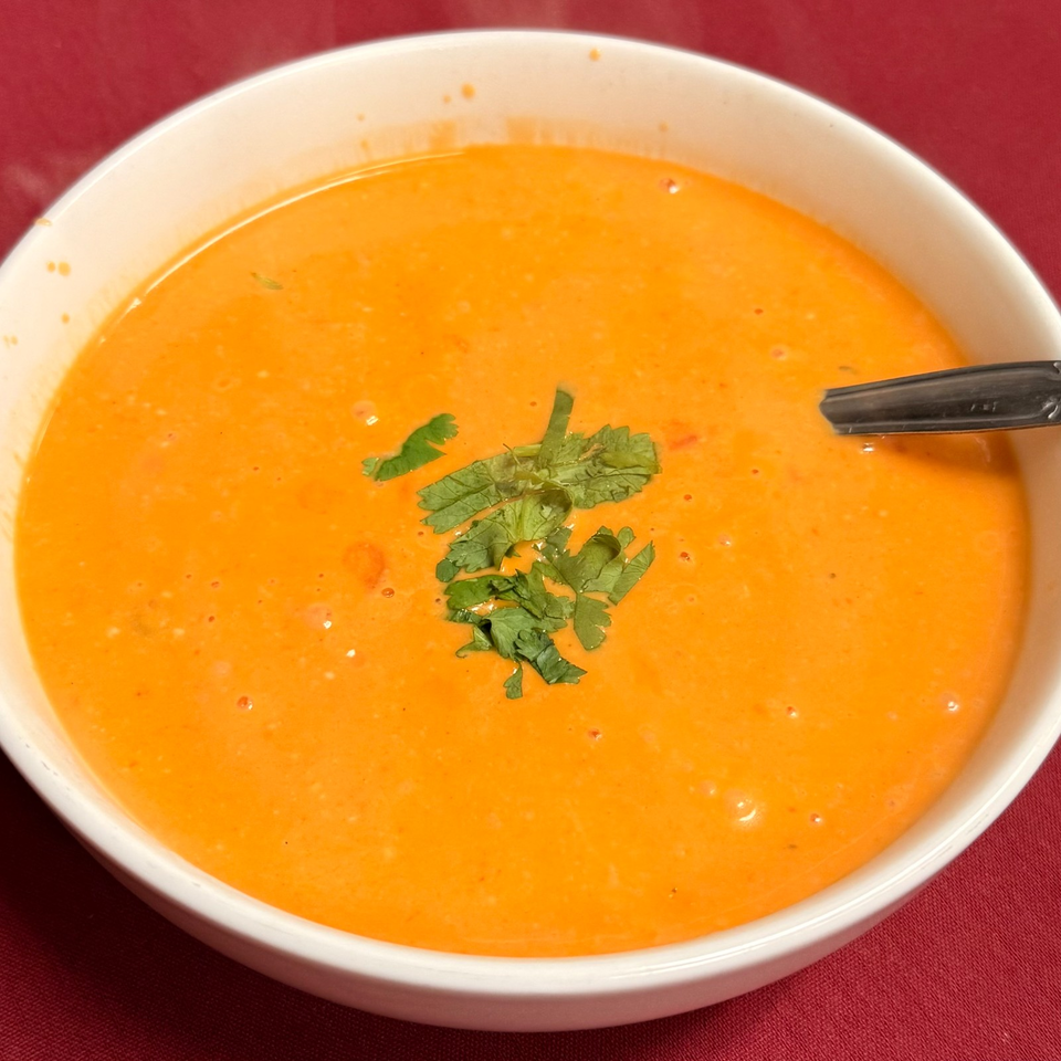 Bowl of creamy orange soup garnished with fresh cilantro, served with a black spoon on a red tablecloth.