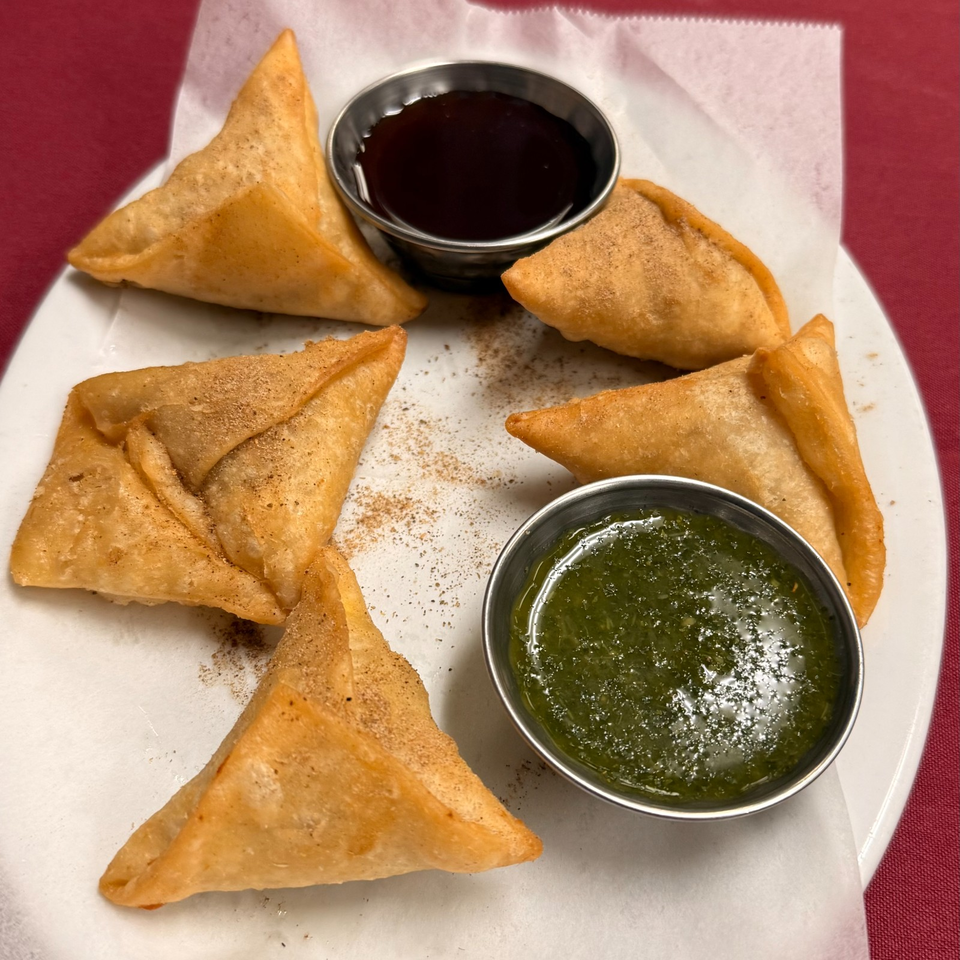 Five golden fried samosas arranged on a plate with bowls of dark tamarind and green herb chutneys.