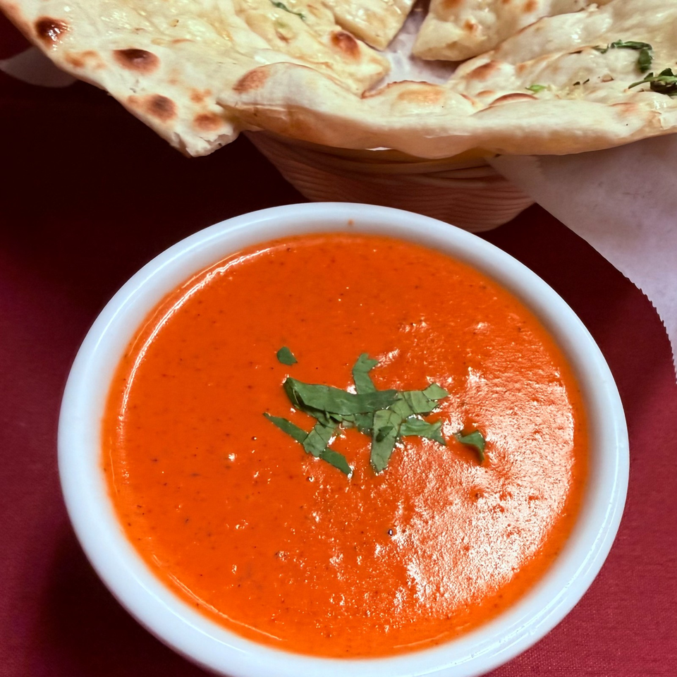 Bowl of creamy orange curry garnished with cilantro, served alongside toasted naan bread on a red tablecloth.