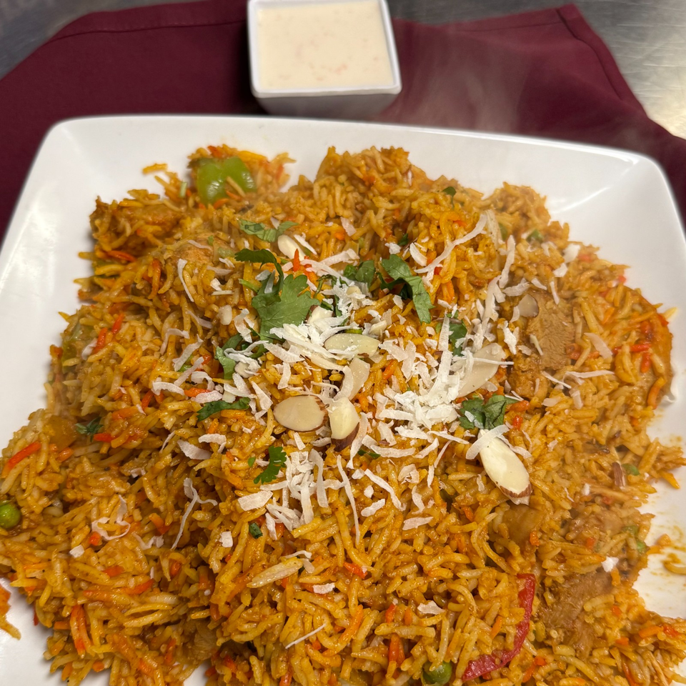 Spiced rice dish topped with shredded coconut, almonds, cilantro, vegetables, and meat, served with creamy dipping sauce.