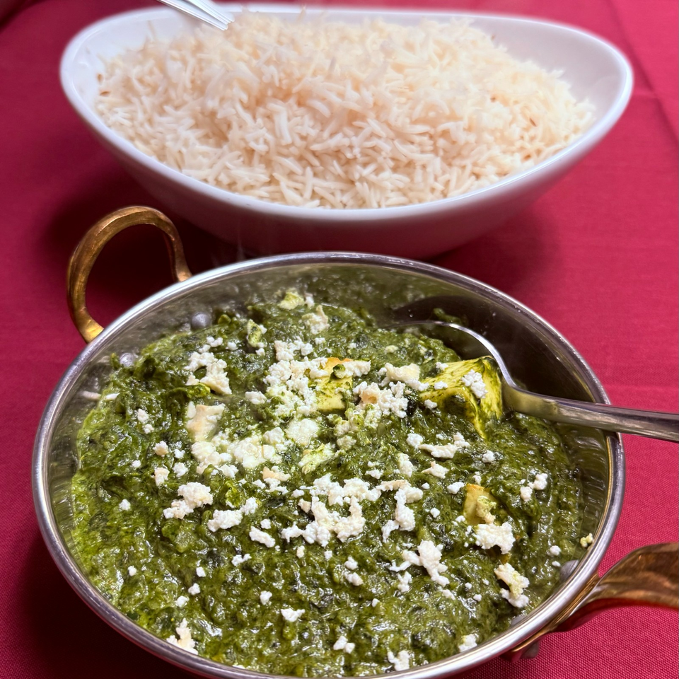Bowl of basmati rice beside creamy spinach curry topped with crumbled cheese, served in metal dishes on a red tablecloth.