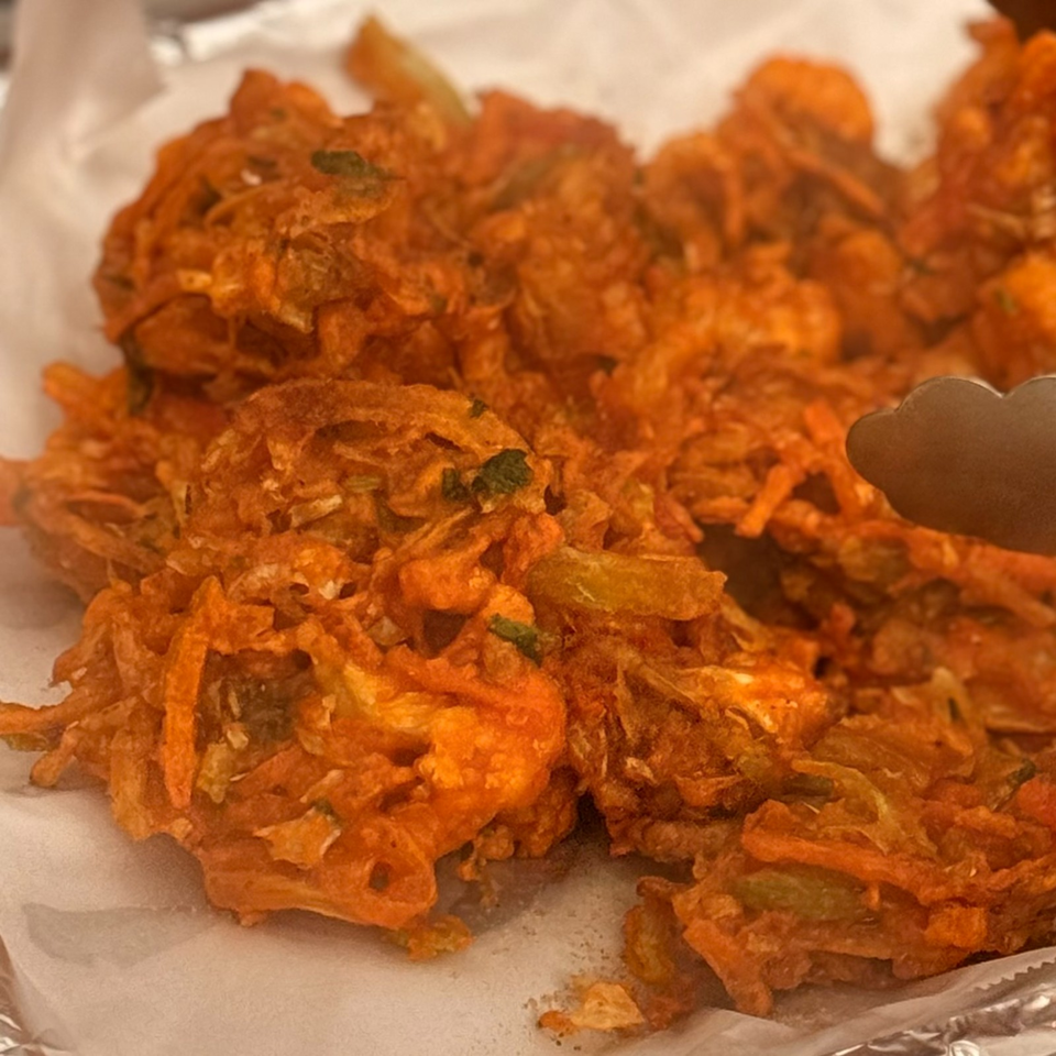Close-up of crispy orange vegetable fritters garnished with green herbs, served on a paper-lined tray with tongs.