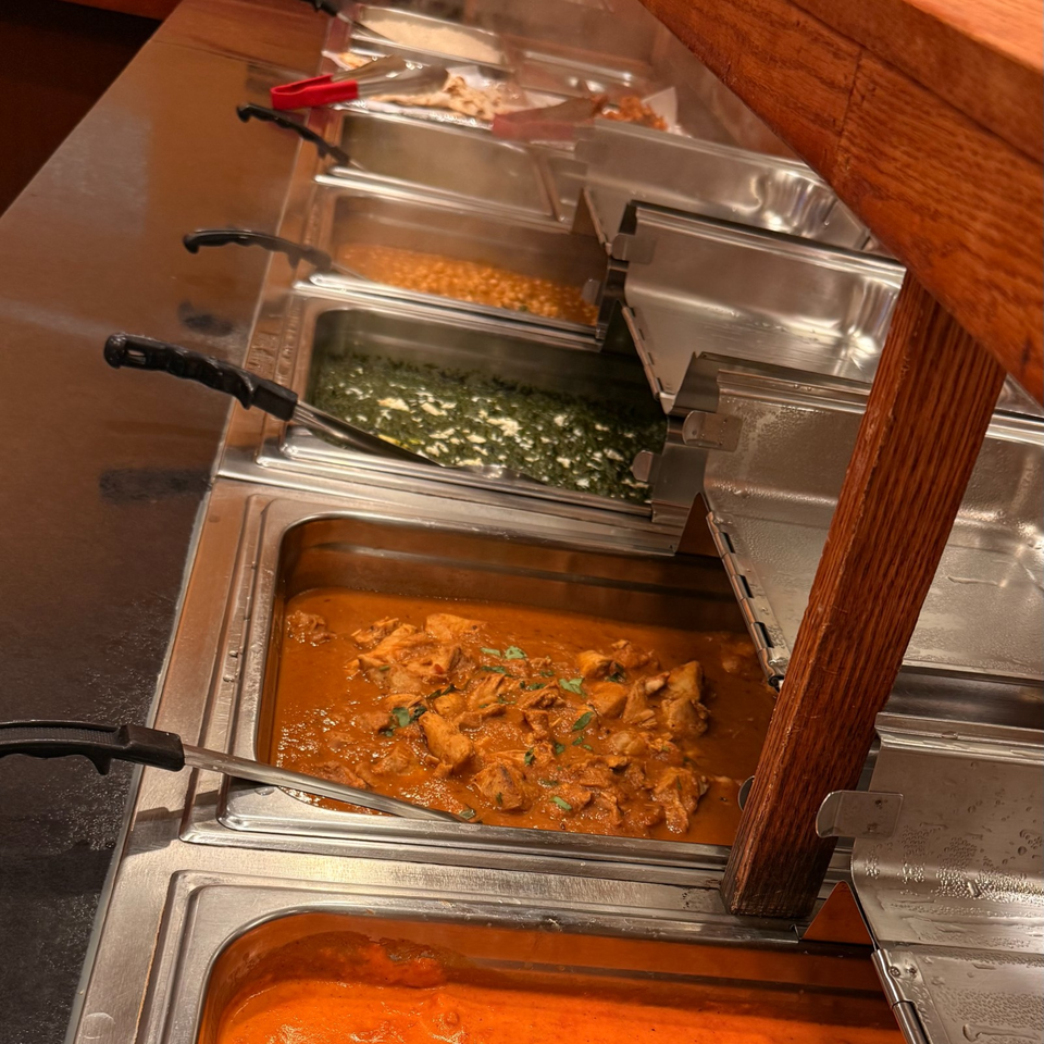 Stainless steel buffet pans hold creamy spinach, chickpeas, and orange chicken curry beneath a wooden sneeze guard.