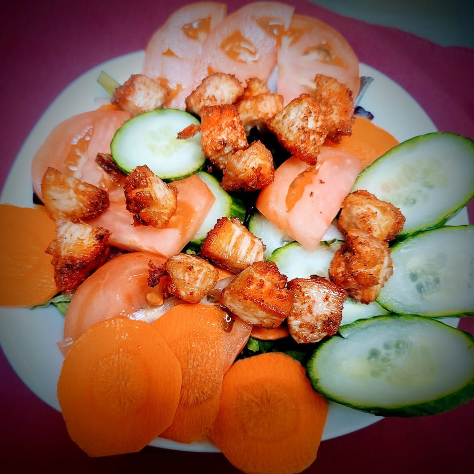 A colorful salad with grilled chicken, sliced tomatoes, cucumbers, and carrots on a white plate.